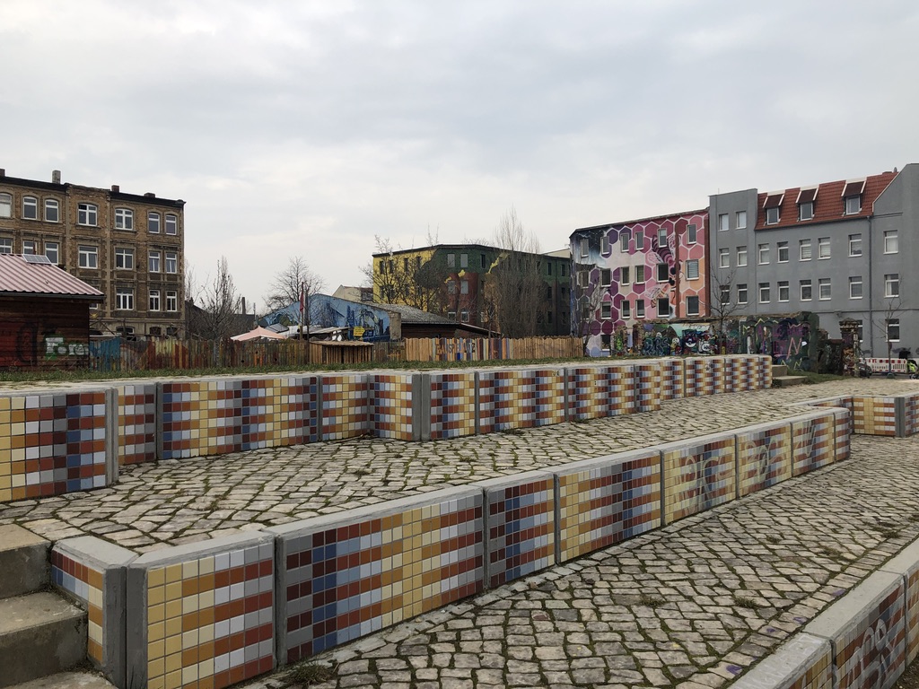 Das Bild zeigt eine Tribüne an einem Sportplatz im Bürgerpark FreiFeld Halle an der Saale. Sie ist mit mehrfarbigen Mosaikfliesen gestaltet. Im Hintergrund sind teilweise mit Streetart gestaltete Häuser des Viertels Halle Freiimfelde sichtbar.