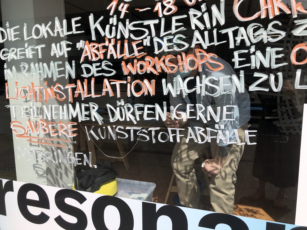 Das Bild zeigt den Ausschnitt einer Fensterscheibe, auf der Fragmente von Sätzen zu lesen sind: Die lokale Künstlerin greift auf "Abfälle" des Alltags, Lichtinstallation, wachsen, Teilnehmer dürfen saubere Kunststoffabfälle mitbringen. Die Scheibe wurde bei einem Projekt der Montag Stiftung Kunst und Gesellschaft aufgenommen, den Reydter Resonanzen.