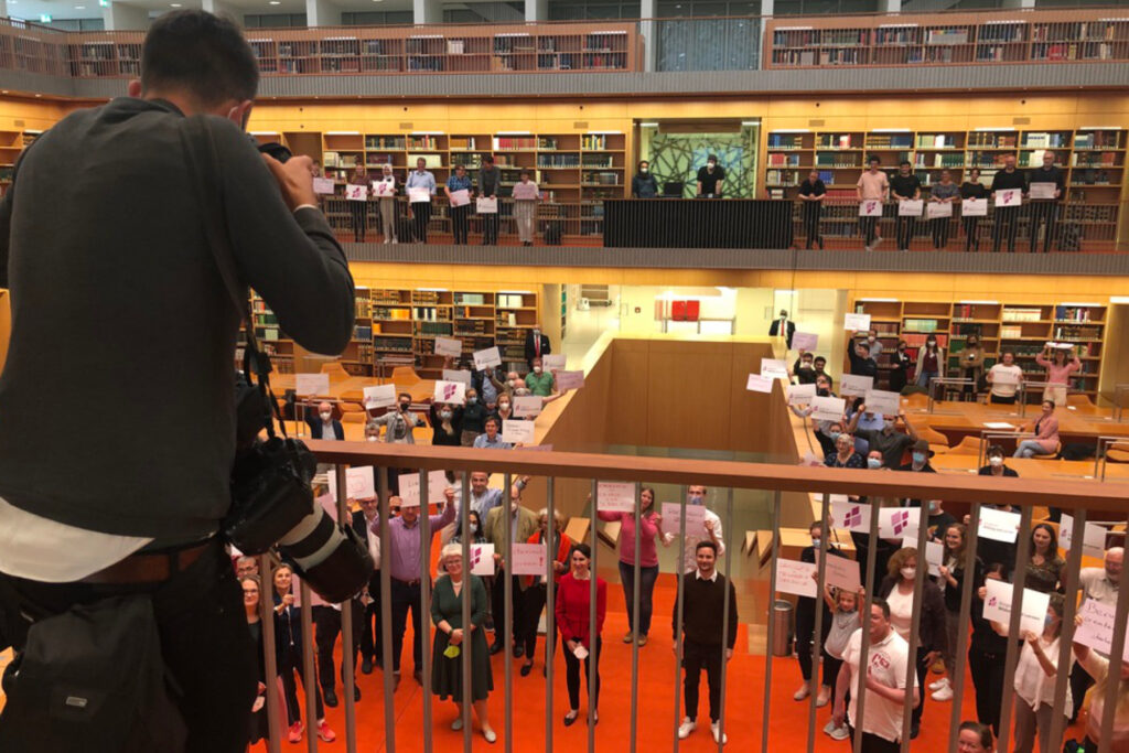 Das Bild zeigt Teilnehmende des Bürgerrats Bildung und Lernen zusammen mit Politikerinnen und Politikerinnen in der Nationalbibliothek Berlin. Von einer Ballustrade herab nimmt ein von hinten sichtbarer Fotograf gerade Bilder von Ihnen auf, während sie ihre Forderungen auf Zetteln in die Kamera halten.