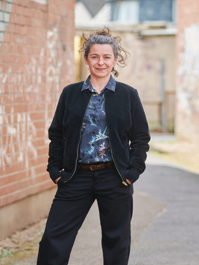 Pauline John steht vor einer mit Graffity-Tags beschriebenen Backsteinwand. Foto: Harald Neumann